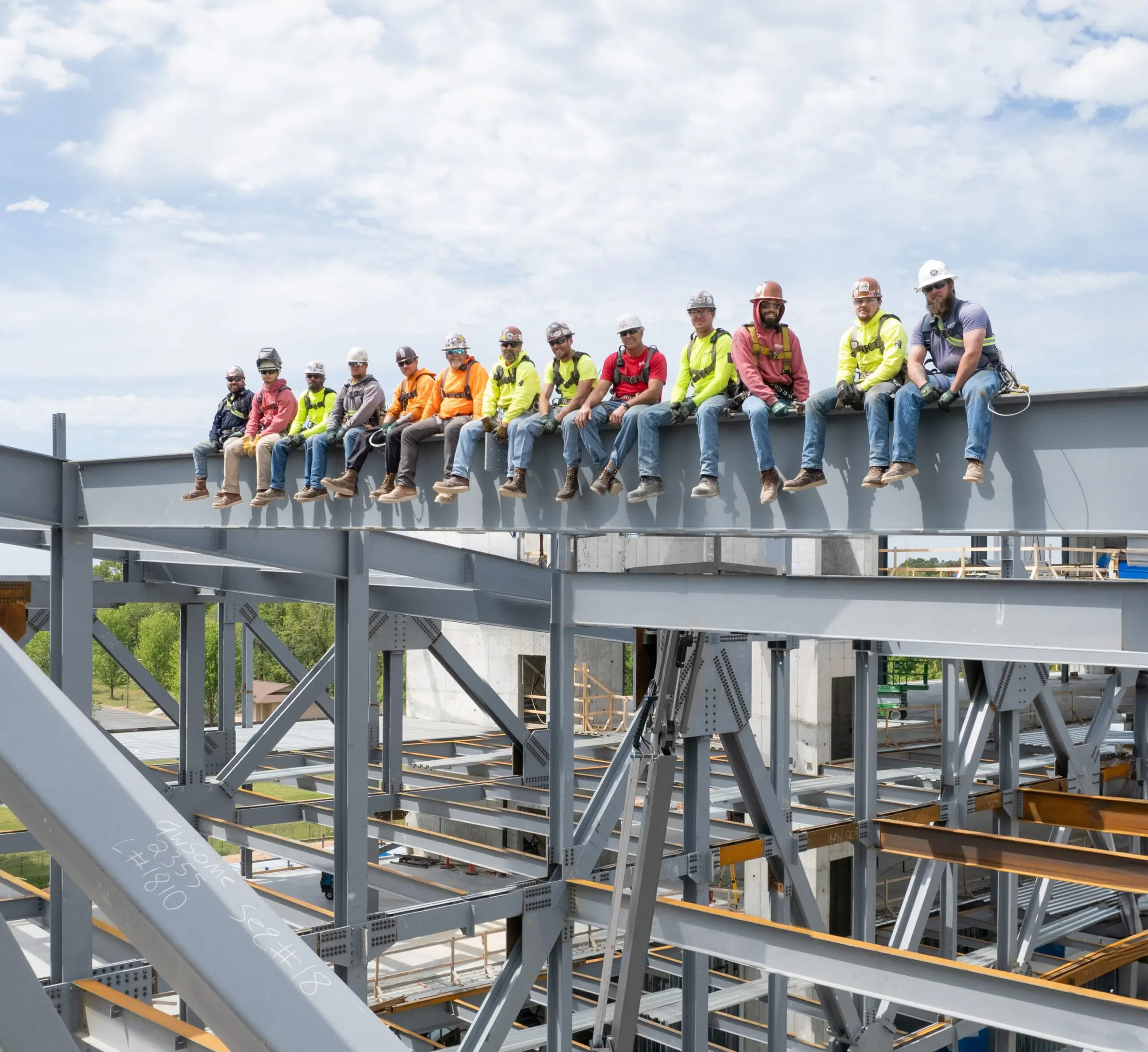 A group of construction workers in safety gear sit side by side on a steel beam high above a building site, with blue sky and clouds in the background.