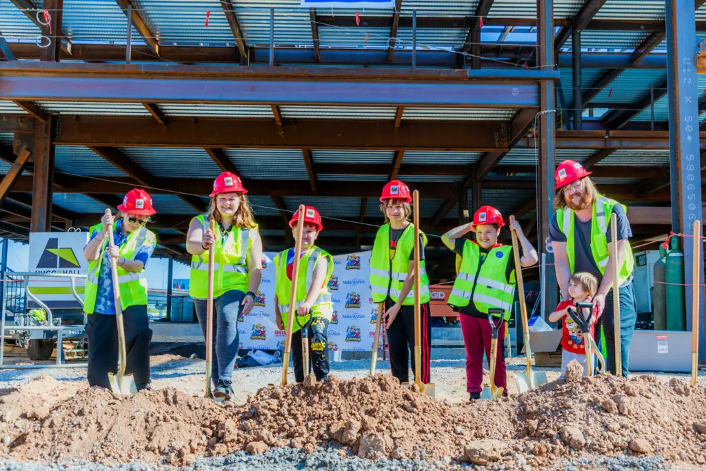 A group of children and one adult, all wearing red hard hats and yellow safety vests, stand in front of a steel building frame, holding shovels at a construction site for a groundbreaking ceremony.