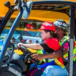 A child wearing a red hard hat and safety vest sits at the wheel of a construction vehicle, guided by an adult in similar safety gear, with a yellow machine in the background.