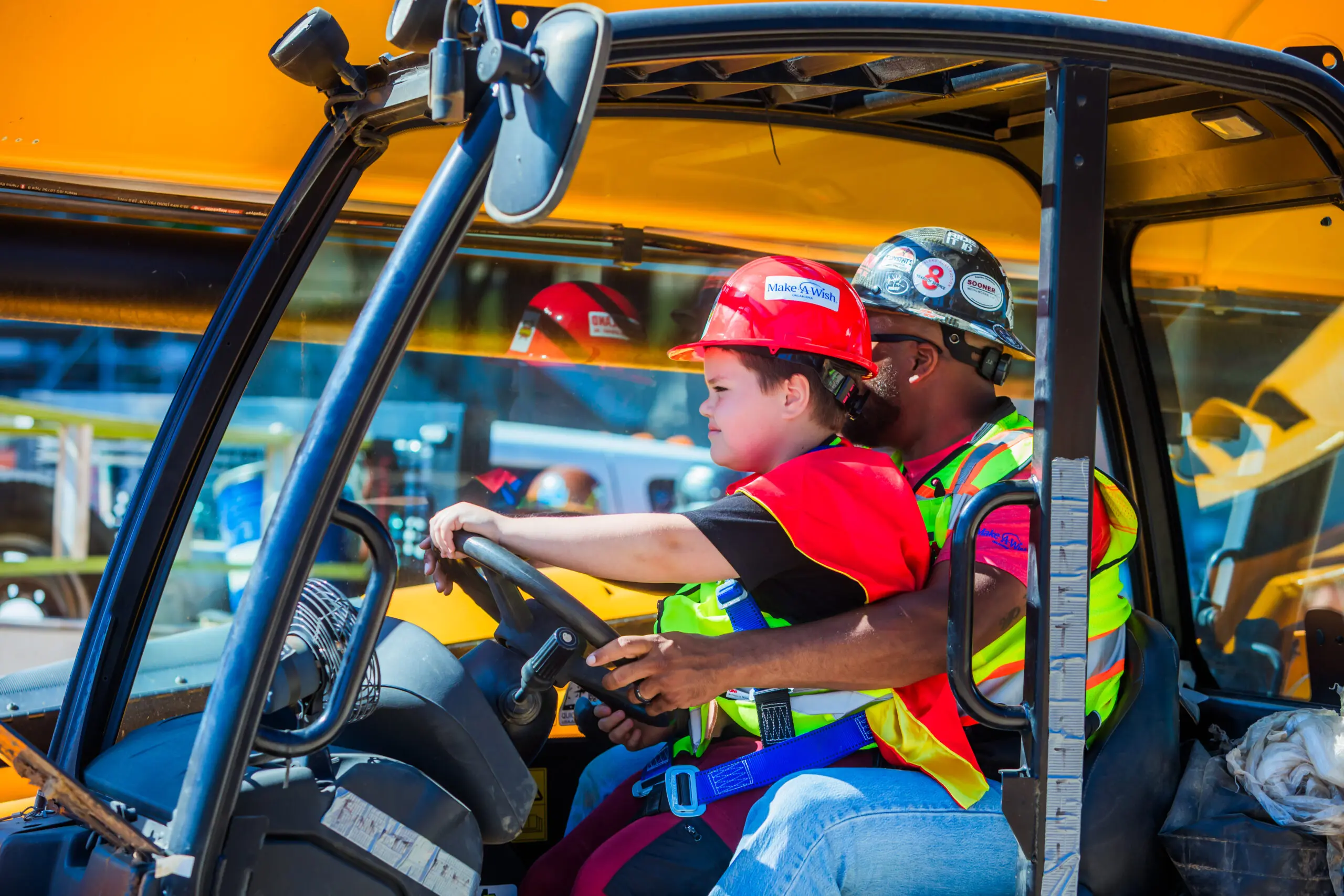 A child wearing a red hard hat and safety vest sits at the wheel of a construction vehicle, guided by an adult in similar safety gear, with a yellow machine in the background.