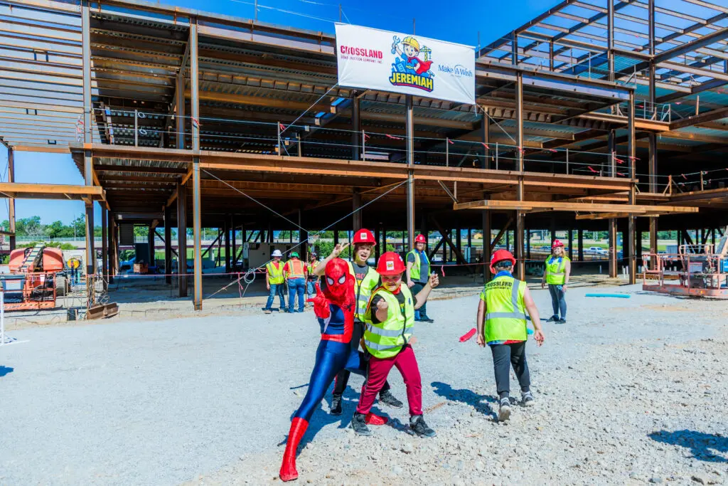 People in safety vests and hard hats pose at a construction site; one person is dressed as Spider-Man. A steel-frame building and a banner with construction company logos are visible in the background.