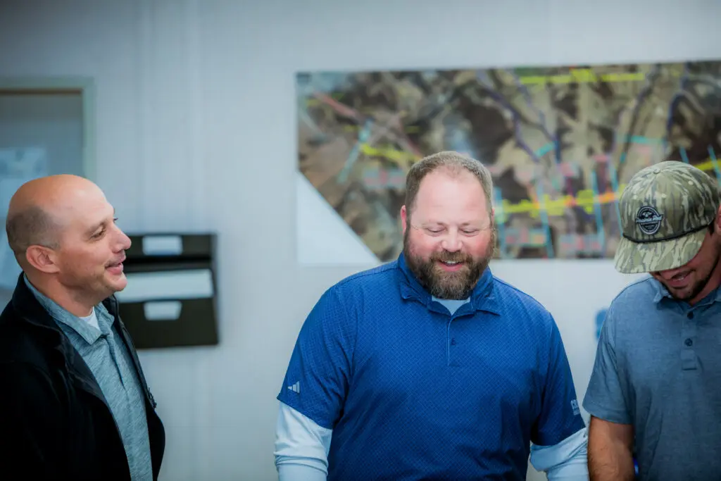 Three men stand together indoors, smiling and talking. A large, colorful map is on the wall behind them. The man in the center wears a blue shirt, while the others wear gray and a camo hat.