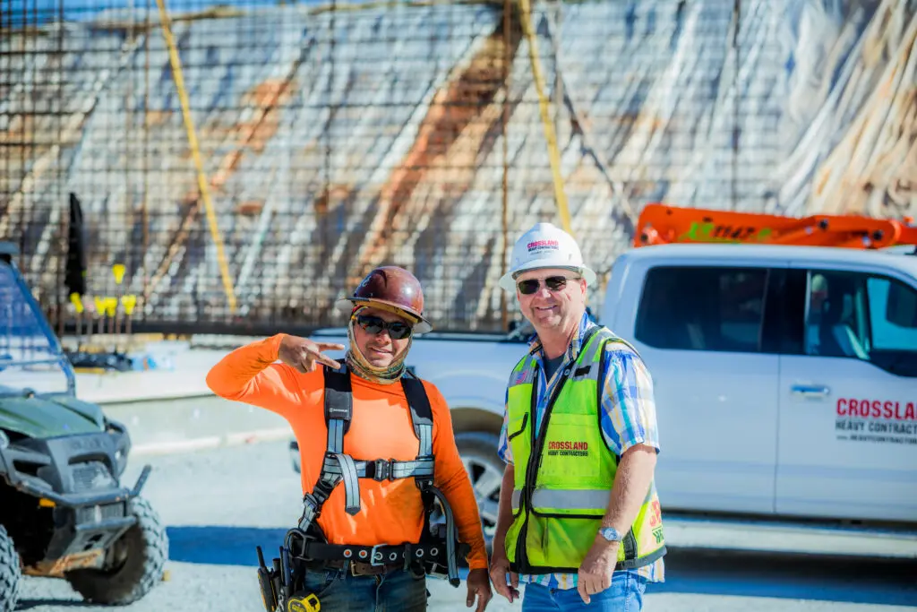 Two construction workers, one wearing an orange shirt and harness, the other in a safety vest and hard hat, stand smiling at a construction site with a white pickup truck and building framework in the background.