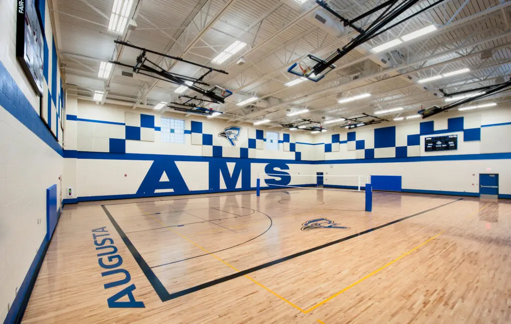 Bright gymnasium with polished wooden floors, blue and white walls, a volleyball net, basketball hoops, large “AMS” letters on the wall, and “AUGUSTA” written on the sideline. School logo appears in several places.
