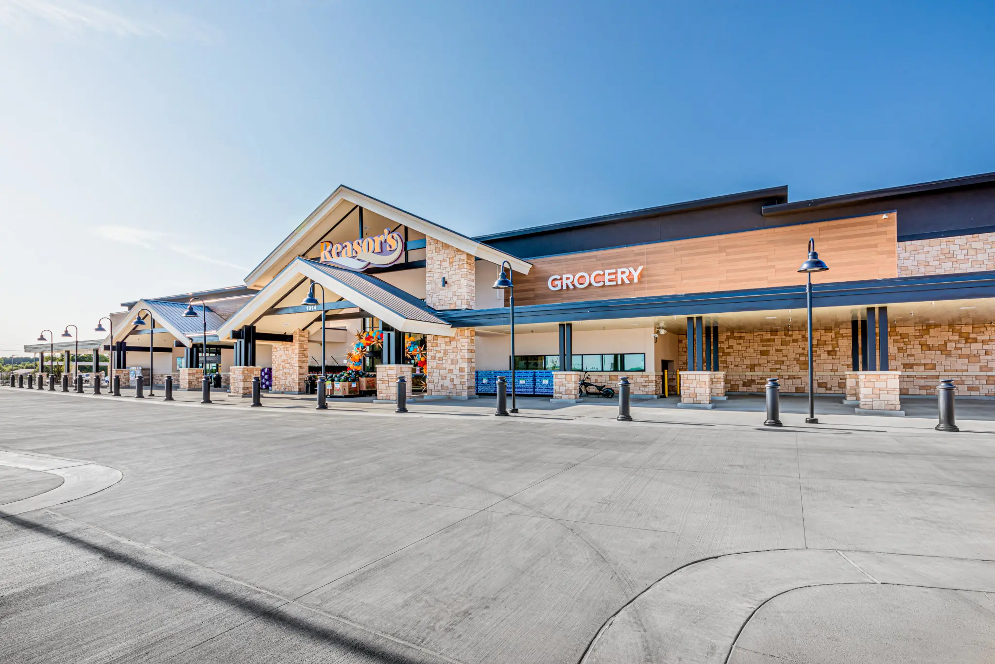 A man in a blue dress shirt and dark blazer smiles while standing outdoors in front of a leafy green tree.