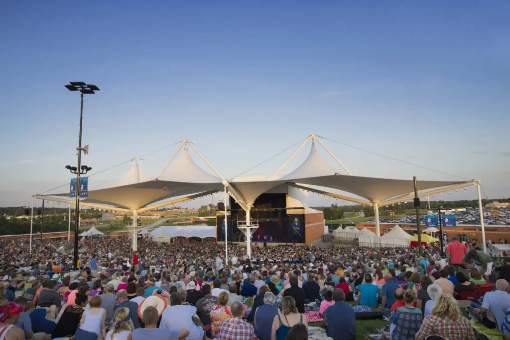 A large crowd sits on a grassy lawn facing an outdoor stage with white canopy roofs at sunset, watching a live concert under a clear blue sky.