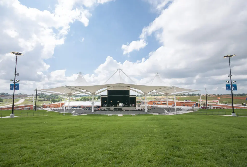 Outdoor amphitheater with a white canopy roof, a large stage, and tiered seating on a grassy lawn under a partly cloudy sky. Surrounding areas feature open land and some buildings in the background.