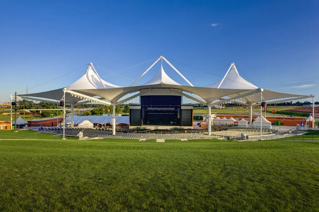 Outdoor amphitheater with a large white canopy roof, open lawn seating in the foreground, rows of chairs in front of the stage, and clear blue sky above.