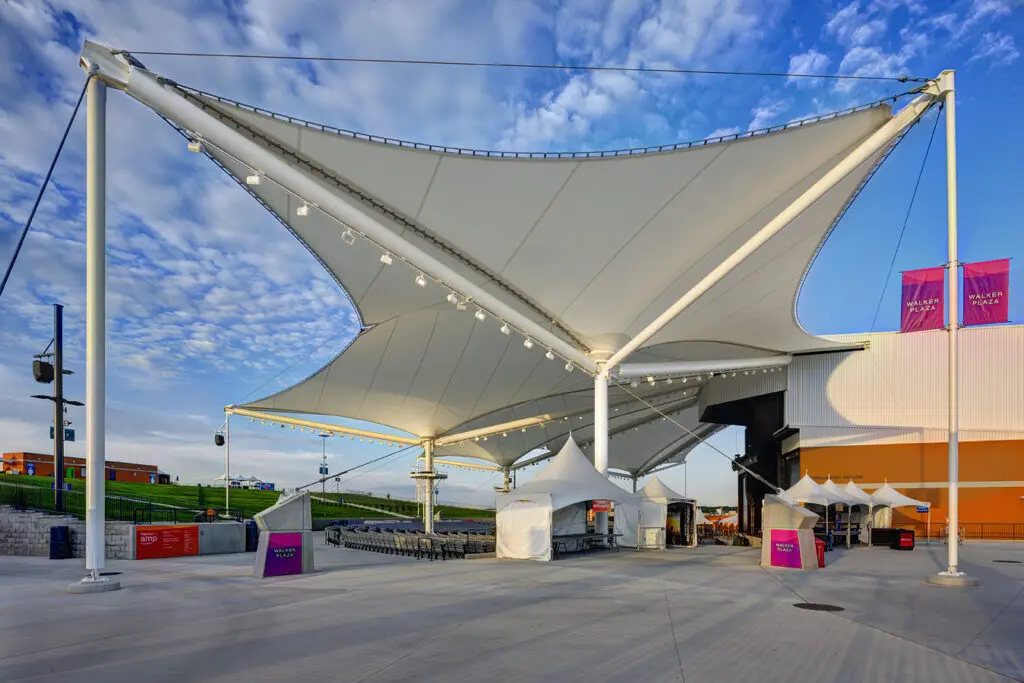 Large white fabric canopy structures with supporting poles provide shade over an outdoor event space. Small tents and chairs are arranged below, with a blue sky and scattered clouds overhead.