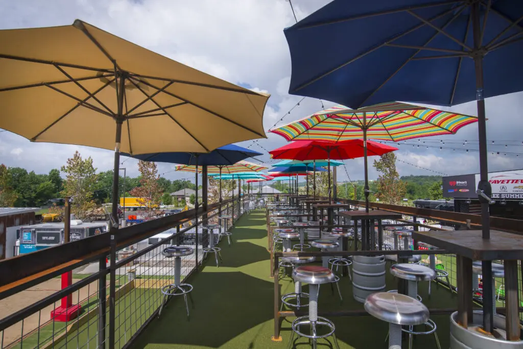 Rooftop patio with metal tables and chairs under large, colorful umbrellas, overlooking a parking lot and trees, with string lights above and a partly cloudy sky in the background.