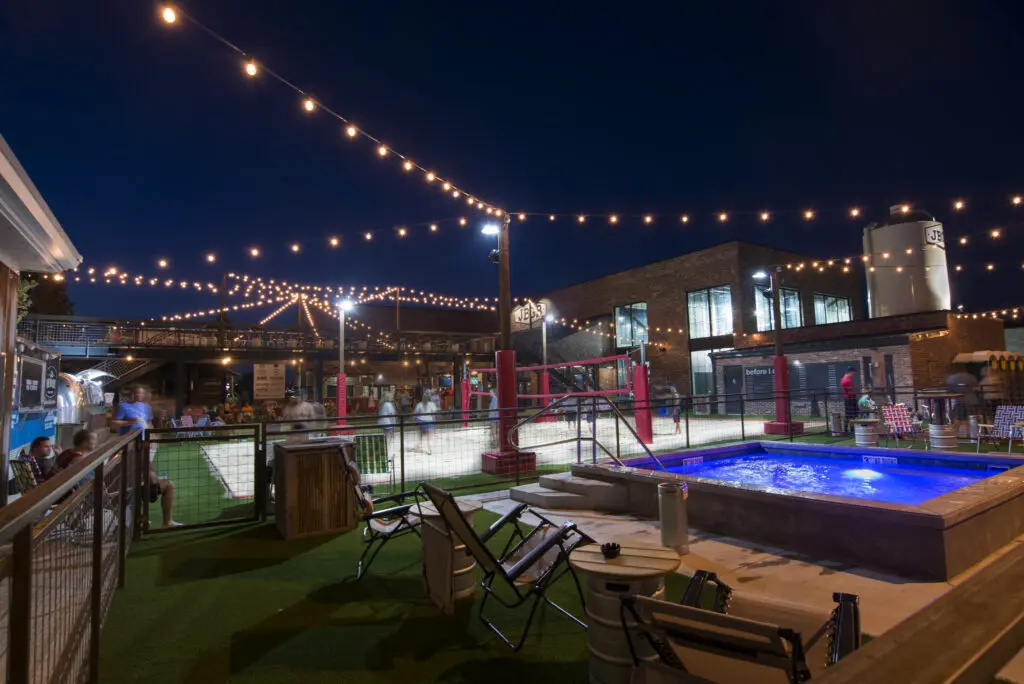 An outdoor venue at night features string lights, a lit hot tub, lounge chairs, and a sand volleyball court. People are socializing in the lively, modern space surrounded by brick buildings.