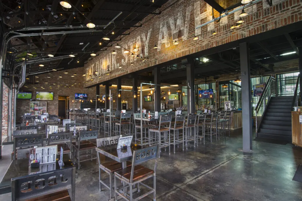 Spacious, industrial-style restaurant with exposed brick walls, metal chairs, high ceilings, and multiple TVs. Large windows and stairs lead to an upper level. Tables are set for dining in a casual, modern atmosphere.