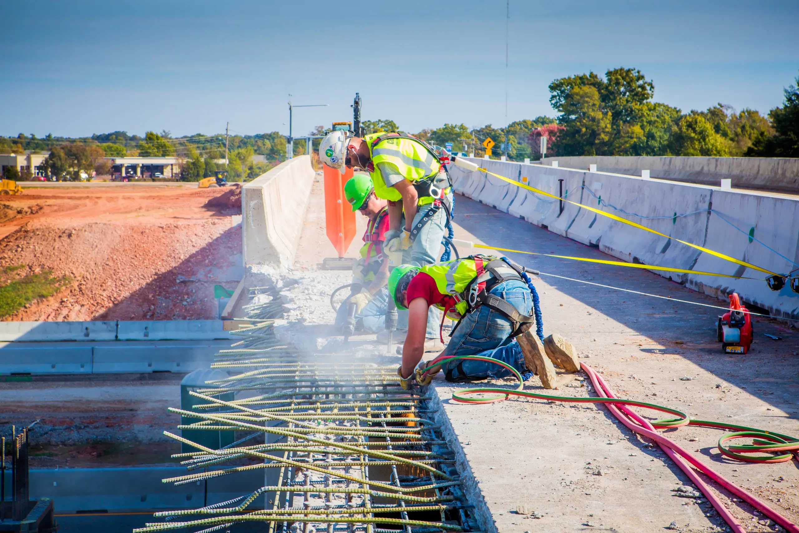 Construction workers in safety gear bend over rebar and concrete on a bridge under construction, using power tools with dust and debris visible; bright safety vests and helmets are worn.