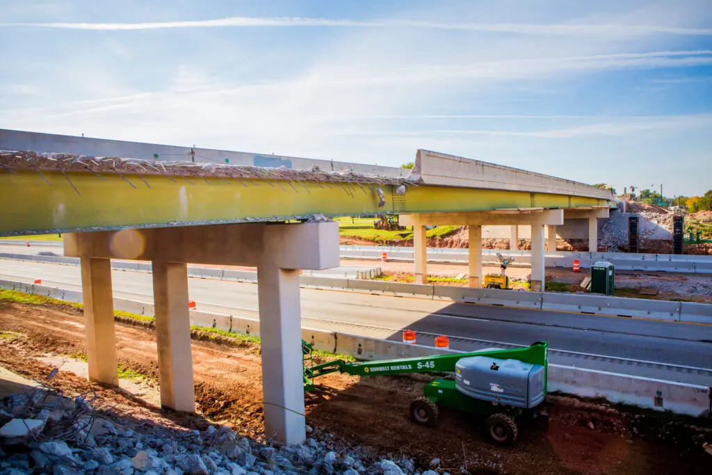 A highway overpass under construction with exposed beams, concrete pillars, and a green construction lift beneath; traffic barriers and equipment are visible on and around the site.