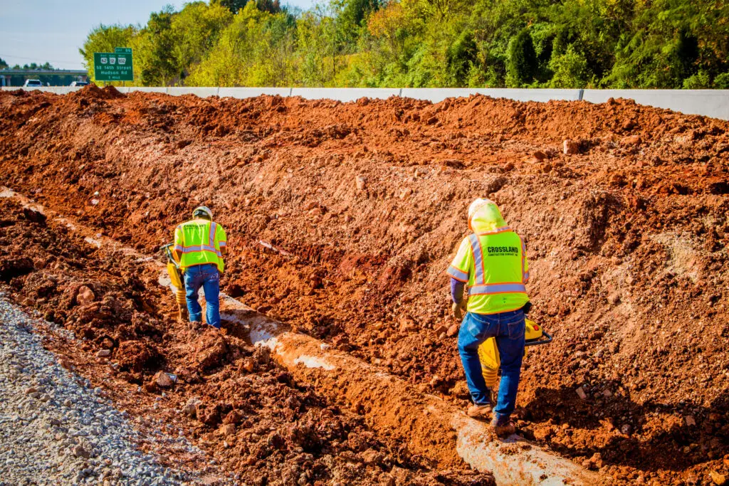 Two construction workers in yellow safety vests work on a muddy roadside, digging or inspecting a trench beside a highway, with trees and a green road sign visible in the background.