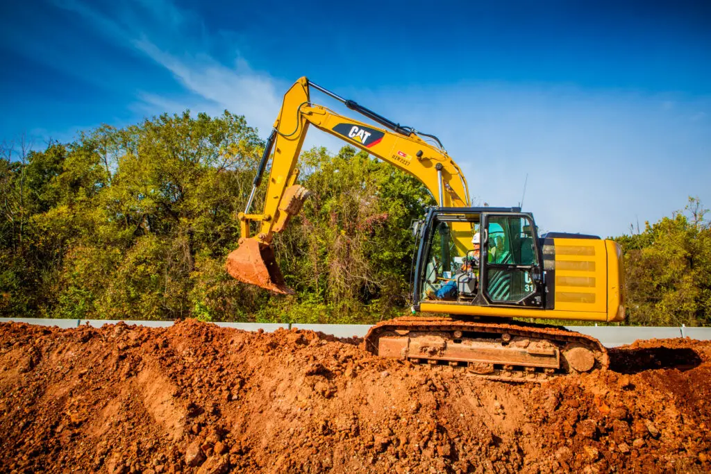 A yellow excavator lifts a bucket of dirt at a construction site, with trees and a clear blue sky in the background.