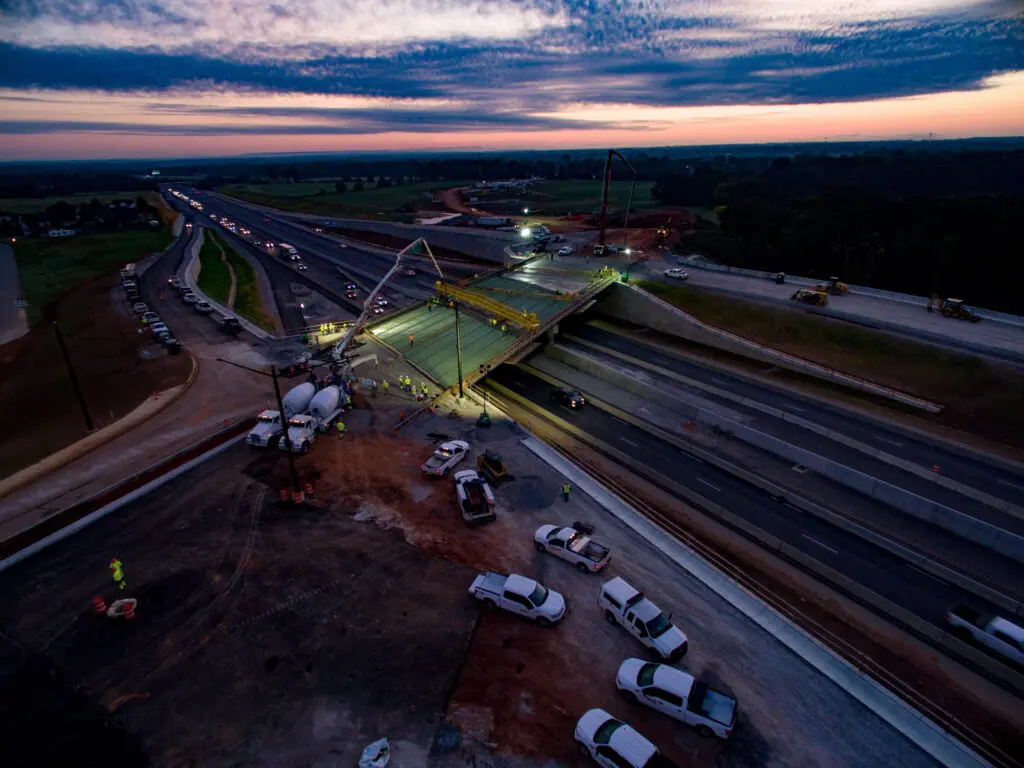 Aerial view of a highway bridge under construction at dusk, with workers, vehicles, and construction equipment on site. Concrete is being poured, and traffic flows on adjacent lanes under a dramatic, cloud-filled sky.