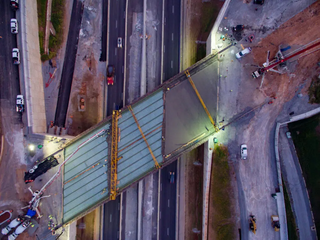 Aerial view of a bridge under construction over a multi-lane road, with vehicles and construction equipment visible, illuminated by work lights in the evening.