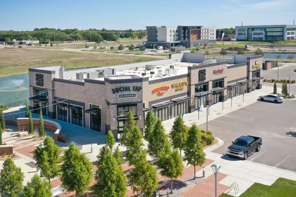 A modern shopping plaza with several restaurants and shops, including Social Tap and Wingz XPRESS. There are a few parked cars and landscaped areas with trees and a fountain in the background.