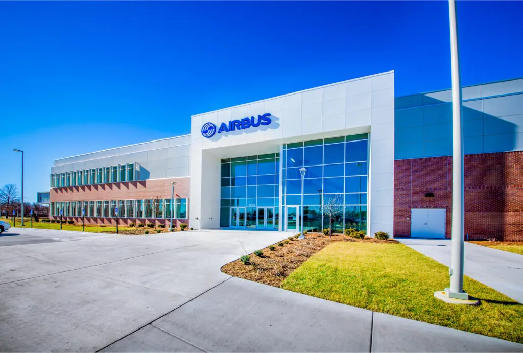 Modern Airbus office building with large glass windows, white and brick exterior, and the Airbus logo above the entrance. Clear blue sky and landscaped grounds with sidewalk and grass are visible in front.