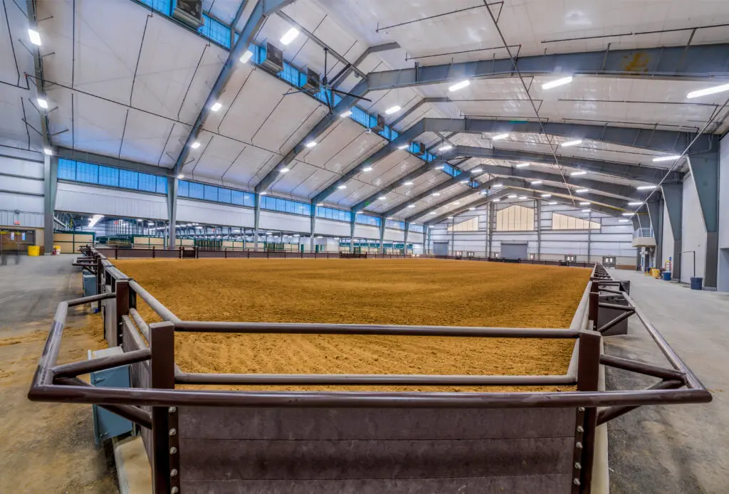 Wide-angle view of an empty indoor rodeo or equestrian arena with a dirt floor, high ceiling, large overhead lights, and steel railings surrounding the arena. The space is spacious and well-lit.