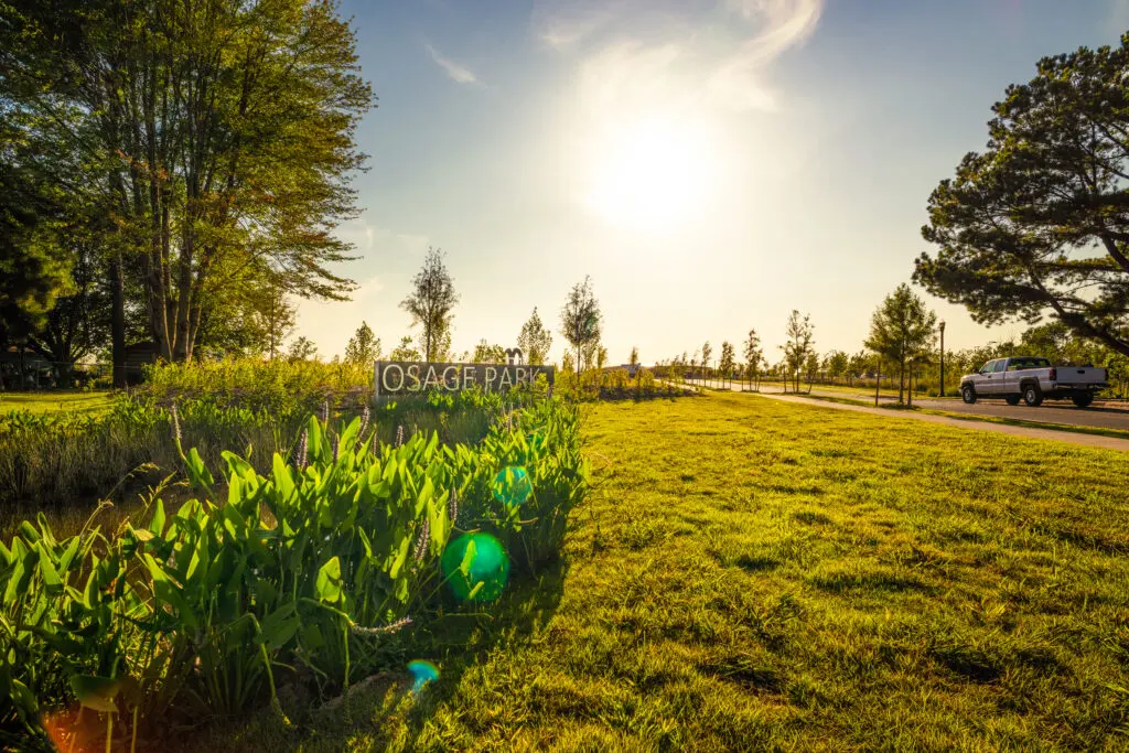 A bright, sunny day at Osage Park with green grass, plants, and trees; a white truck is parked on a road to the right, and the sun is shining through the sky, creating lens flares.