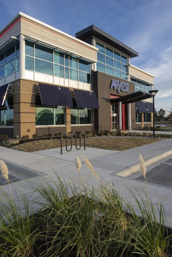 A modern bank building with large windows, blue awnings, and an "ARVEST" sign above the entrance. A bike rack and ornamental grasses are in the landscaped area near the sidewalk.