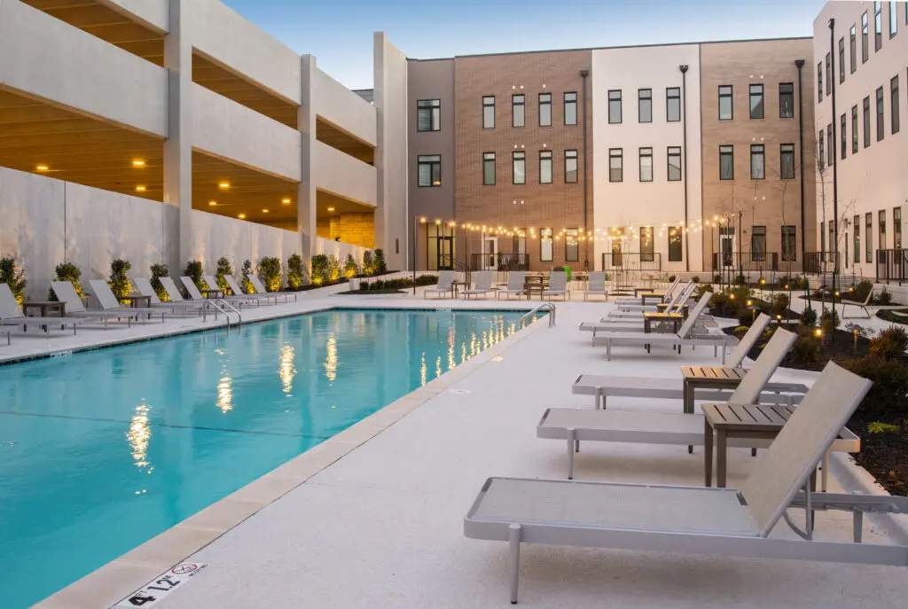 Modern outdoor swimming pool area with lounge chairs lined up along the sides, string lights hanging overhead, and a multi-story apartment building and parking garage in the background at sunset.