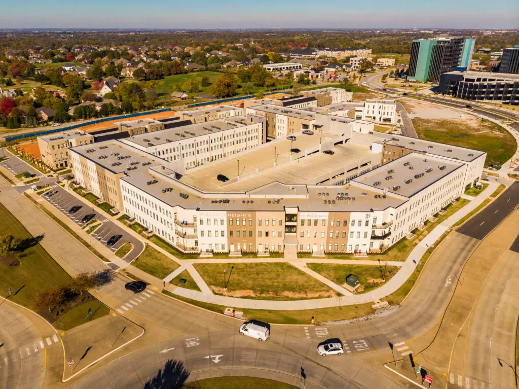 Aerial view of a large, modern, U-shaped apartment complex with surrounding roads and parked cars, set in a suburban area with houses, trees, and nearby office buildings.