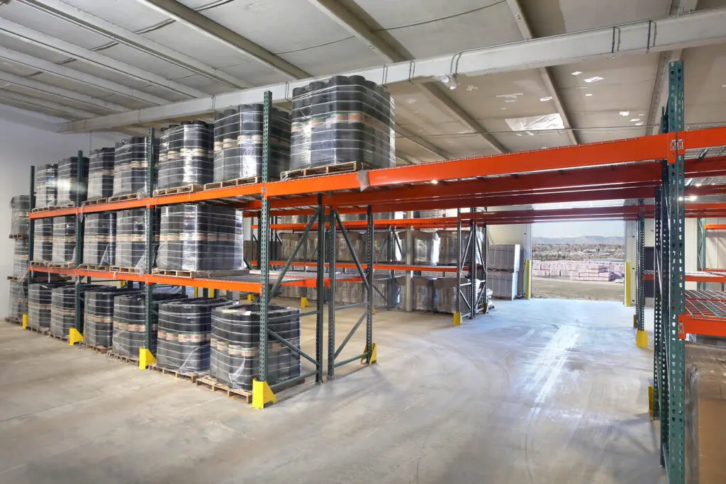 Large industrial warehouse with tall metal shelves holding stacks of coiled wire or cable. Sunlight streams in through an open door, revealing rows of coils and a view of an outdoor area beyond. The floor is clean and spacious.