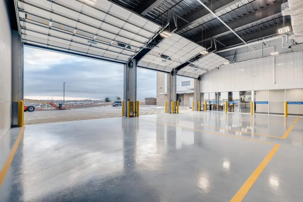 Spacious, clean industrial garage with shiny gray floors, high ceilings, and large open bay doors. Yellow lines mark lanes on the floor, and glass doors lead to an interior area. Outdoor view shows a cloudy sky and some snow.