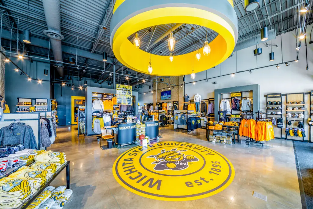 A brightly lit Wichita State University merchandise store, featuring school apparel, hats, and accessories. The floor displays a large yellow WSU logo, and yellow accents decorate the modern space.