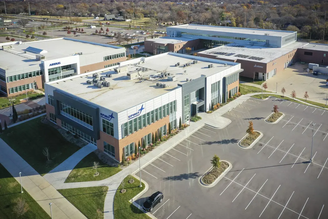 Aerial view of a modern office complex with several large brick and glass buildings, parking lots, and landscaped areas. Only a few cars are parked, and autumn trees line the lots and roads.