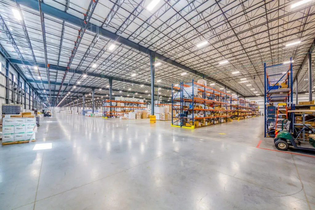 Spacious warehouse interior with tall metal shelves holding boxes and goods, bright overhead lighting, and wide polished concrete floors, creating an organized and orderly environment.