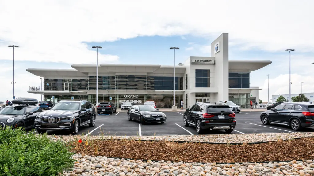 A modern BMW dealership with large glass windows, a "Grand Reopening" sign, and several parked cars in front. The sky is partly cloudy and landscaping with rocks and mulch is visible in the foreground.