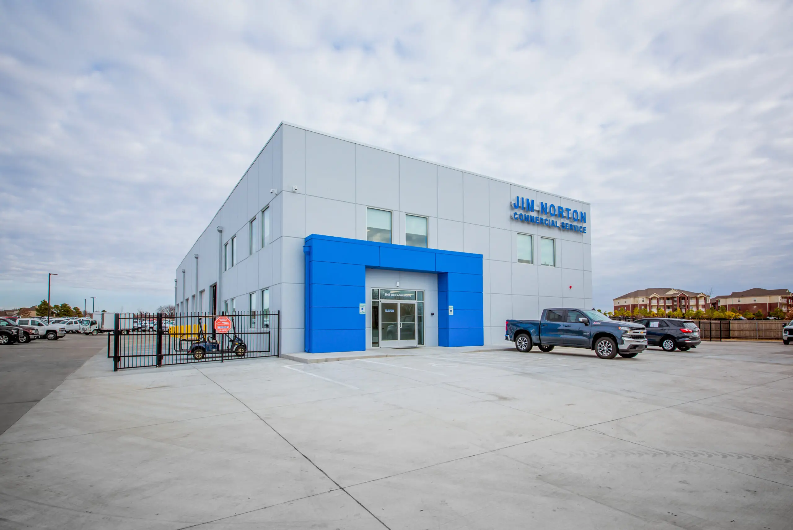 A modern, two-story commercial building with gray and blue precast concrete accents, labeled "Jim Norton." Several cars are parked nearby under a cloudy sky. The area is spacious with a gated section to the left.