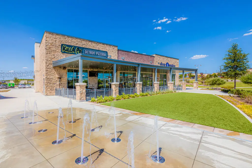 A modern restaurant with a stone exterior and large glass windows, featuring outdoor seating and a patio. Water fountains spray from the pavement in front, and there is a neatly maintained lawn nearby under a clear blue sky.