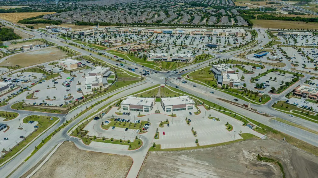 Aerial view of a suburban intersection with multiple shopping centers, large parking lots, and grid-patterned roads. Residential neighborhoods are visible in the background.