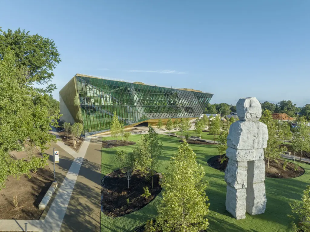A modern, glass-walled building stands behind a large stone sculpture in a landscaped park with green grass, young trees, and walking paths under a clear blue sky.