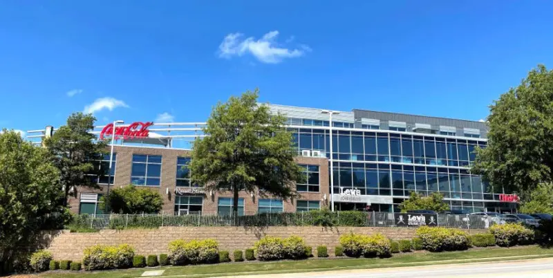 A modern office building with large glass windows and a Coca-Cola sign on top, surrounded by trees, shrubs, and a street in the foreground under a blue sky.