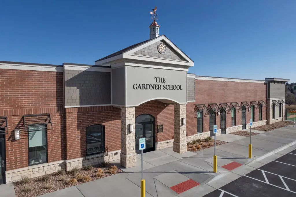 A modern brick building with a sign reading "The Gardner School" above the entrance, featuring large windows, a clock tower, and an accessible parking area in front under a clear blue sky.