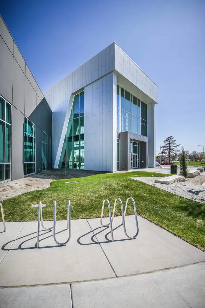 A modern building with large glass windows and metal panels, surrounded by green grass under a clear blue sky; empty metal bike racks are in the foreground on a concrete path.
