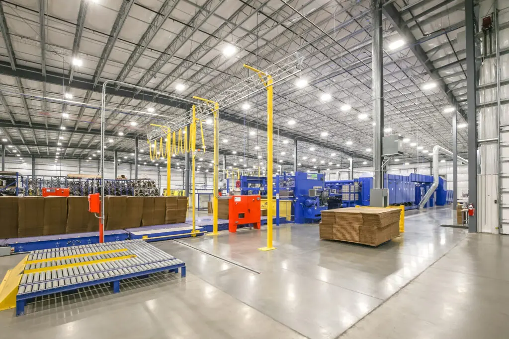 A spacious, well-lit warehouse with stacks of cardboard boxes, conveyor belts, and large blue and yellow industrial machinery, all organized on a polished concrete floor under a high metal ceiling.