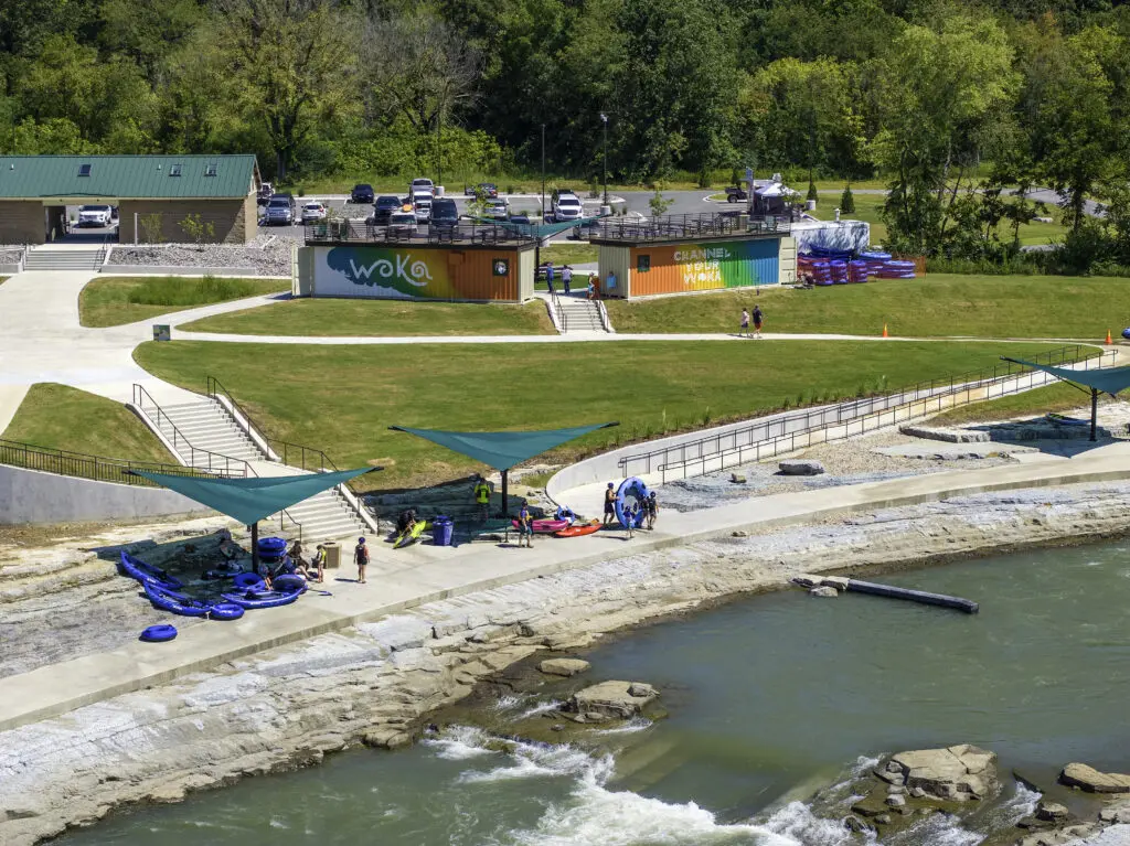 A riverside recreation area features grassy lawns, a modern building with "WOKA" signage, parked cars, and people preparing for water activities near blue rafts and sun shades by a flowing river.