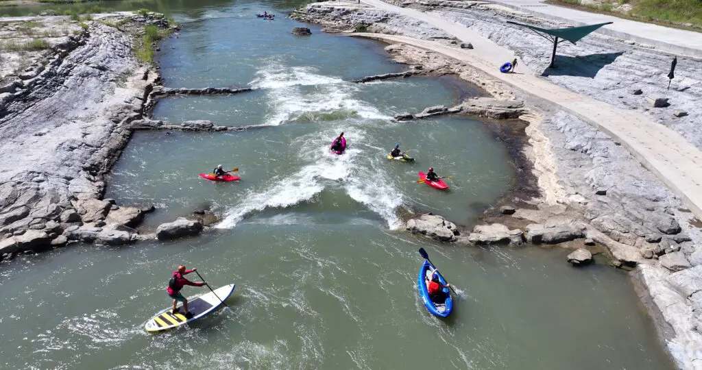Several people are kayaking and paddleboarding on a river with rocky banks, navigating through small rapids. The river is bordered by stone and a walking path, with one person sitting under a shaded area.