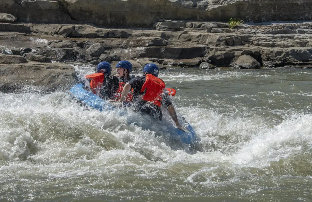 Three people in blue helmets and orange life jackets are white-water rafting through rough rapids, with rocky terrain and flowing water surrounding them.