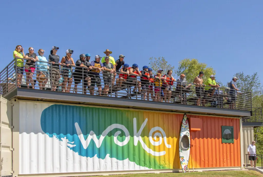 A group of people stand on a rooftop deck above a colorful shipping container painted with a wave and the word "WOKA," while a kayak leans against the container and a person stands alone below. The sky is clear and blue.