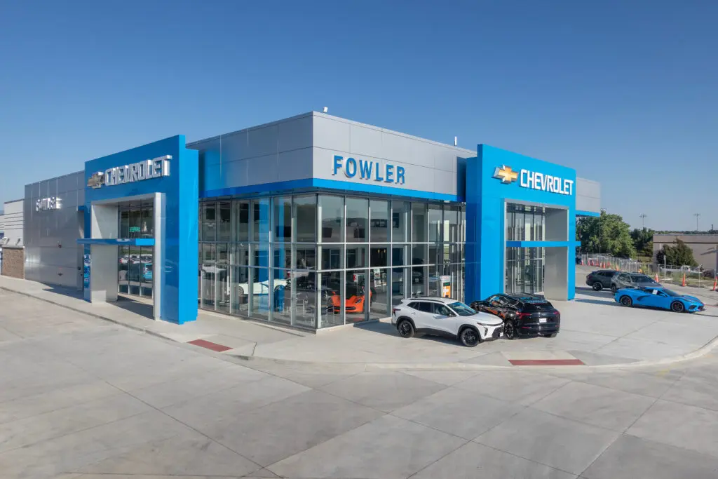 Modern Chevrolet dealership building with large glass windows, blue accents, and "Fowler" and "Chevrolet" signs. Several cars, including black and white models, are parked in front under a clear blue sky.