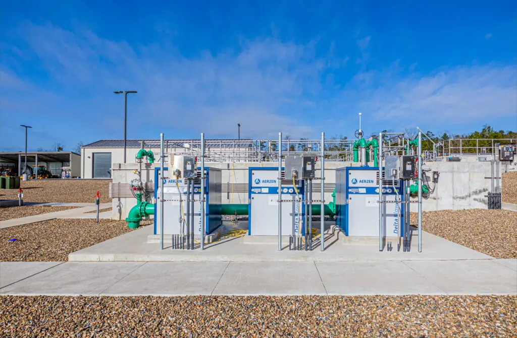 Outdoor water treatment facility with three large filtration units, green pipes, and control equipment on a gravel-covered ground under a clear blue sky. A building and additional infrastructure are in the background.