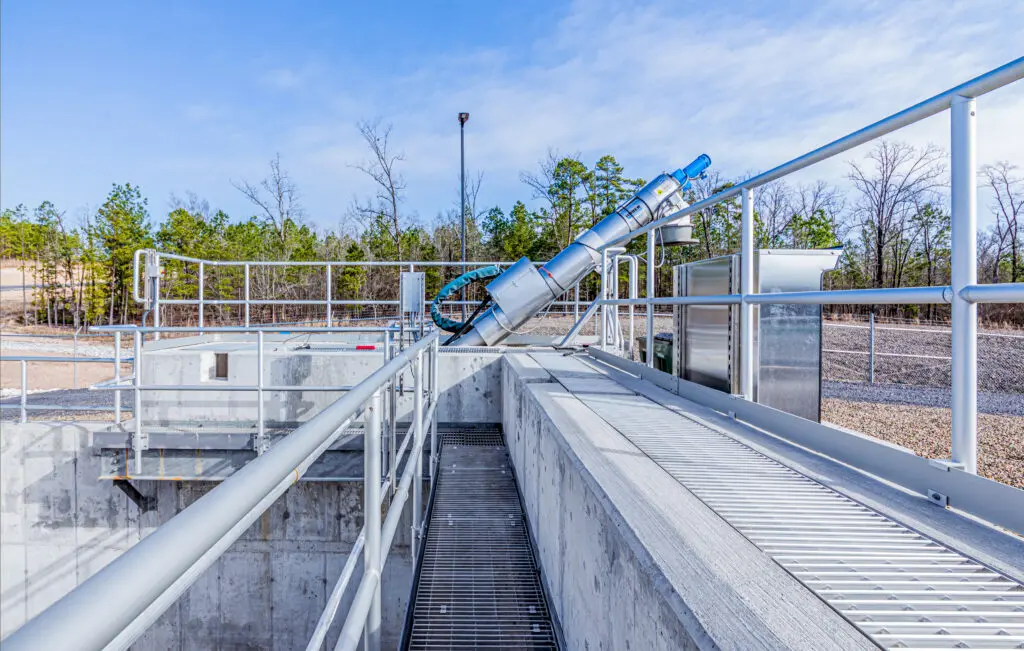 A modern outdoor water treatment facility features metal railings, walkways, and a large inclined mechanical device, with trees and a blue sky visible in the background.
